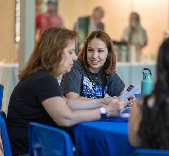 Mother reading a pamphlet and smiling daughter speaking to her while both sitting at franciscan center table during a prospective students info session